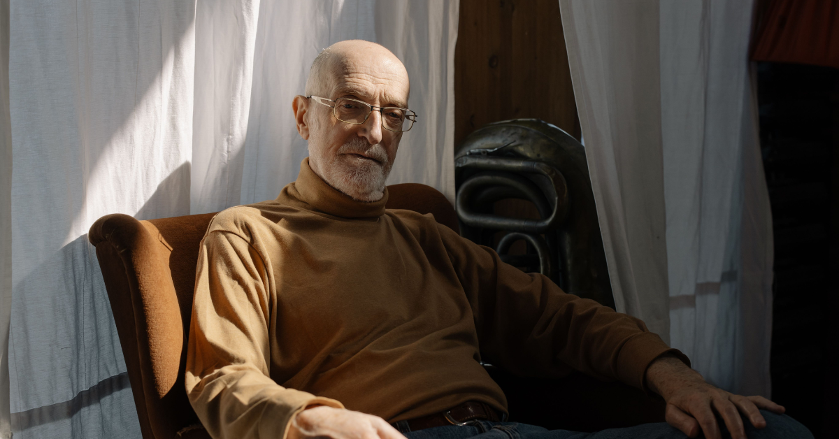 An older adult sitting comfortably in an armchair, looking thoughtful in a softly lit room with natural daylight.